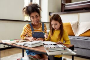 Caucasian little girl spending time with african american baby sitter. They are drawing, learning to write letters, reading a book. Children education, leisure activities, babysitting concept